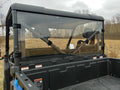 A rear view of a utility vehicle fitted with the Cfmoto Uforce 1000 Lexan Back Panel, featuring an empty black cargo bed, clear rear windshield, and headrests. Its parked in a grassy field with leafless trees under a cloudy sky; safety stickers visible.