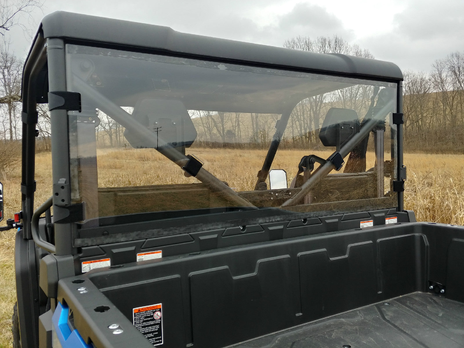A rear view of a utility vehicle fitted with the Cfmoto Uforce 1000 Lexan Back Panel, featuring an empty black cargo bed, clear rear windshield, and headrests. Its parked in a grassy field with leafless trees under a cloudy sky; safety stickers visible.