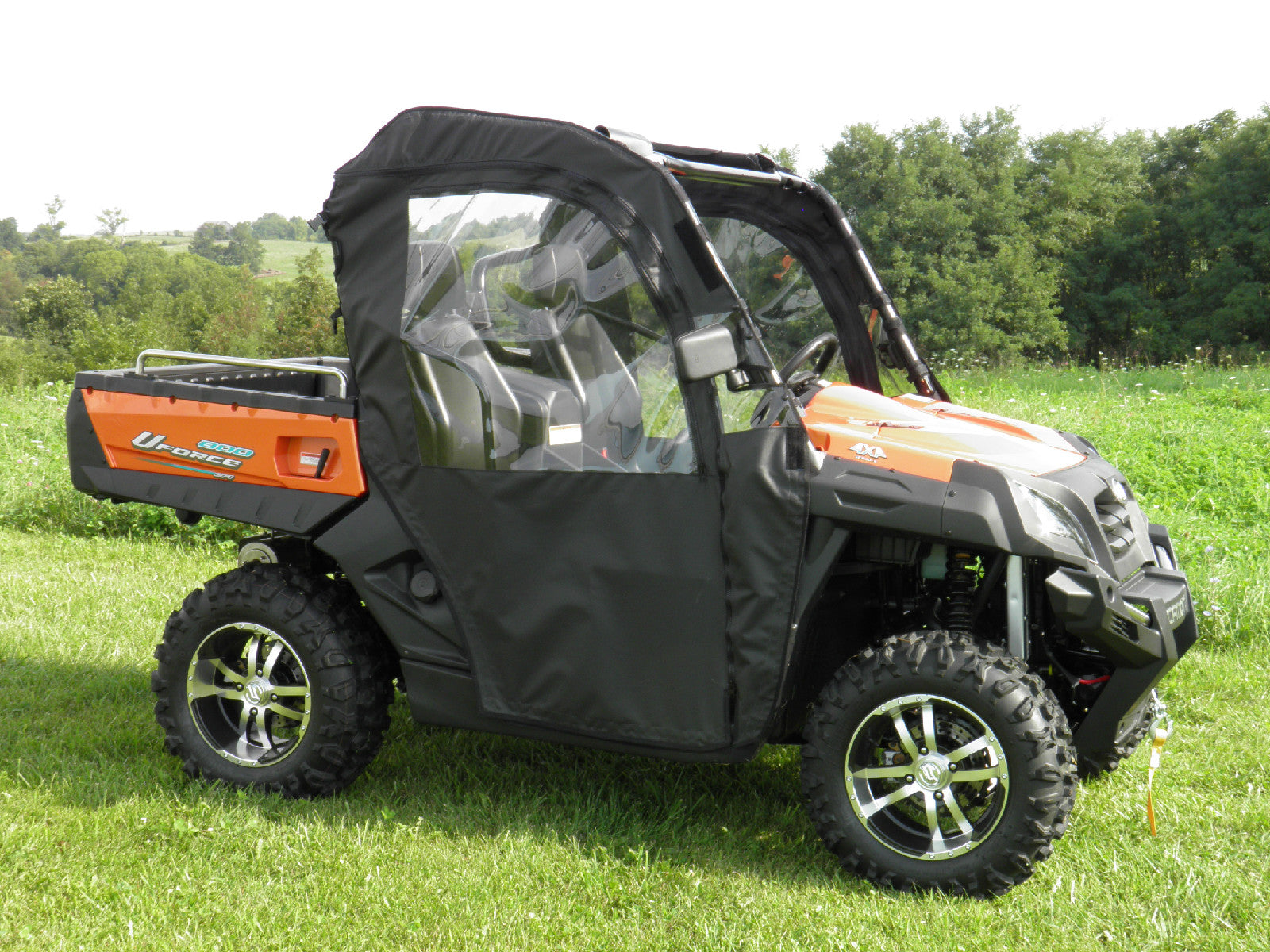 A Cf Moto Uforce 500/800 Soft Doors Kit with clear plastic windows is installed on a black and orange utility vehicle sitting on green grass, with trees and a cloudy sky in the background.