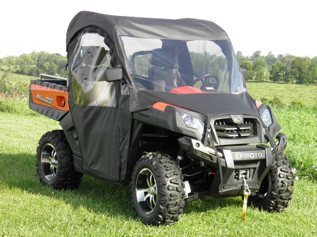 A black and orange CFMOTO UTV with off-road tires features a Cf Moto Uforce 500/800 Soft Full Cab Enclosure With Vinyl Windshield, parked on green grass. It has a front winch, clear vinyl windows, and sits under a bright sky with trees in the background.