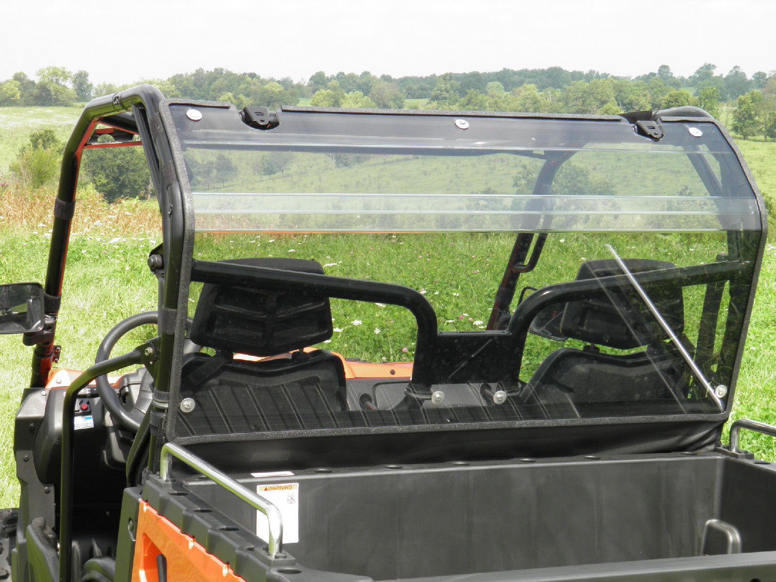 A Cfmoto Uforce 500/800 2pc. Lexan Back Window is installed on an orange utility vehicle in a grassy field, with the window partially open and two black seats inside. Trees and a cloudy sky are visible in the background.