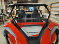 A red CFMOTO Zforce 950 with a half windshield, two bucket seats, and black roll bars is parked indoors. The front features visible branding, with workshop shelves, a large fan, and equipment in the background.