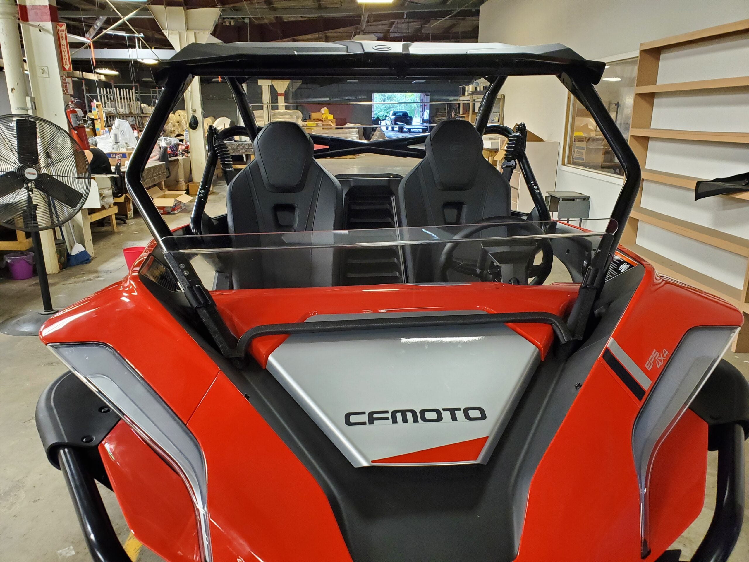 A red CFMOTO Zforce 950 with a half windshield, two bucket seats, and black roll bars is parked indoors. The front features visible branding, with workshop shelves, a large fan, and equipment in the background.