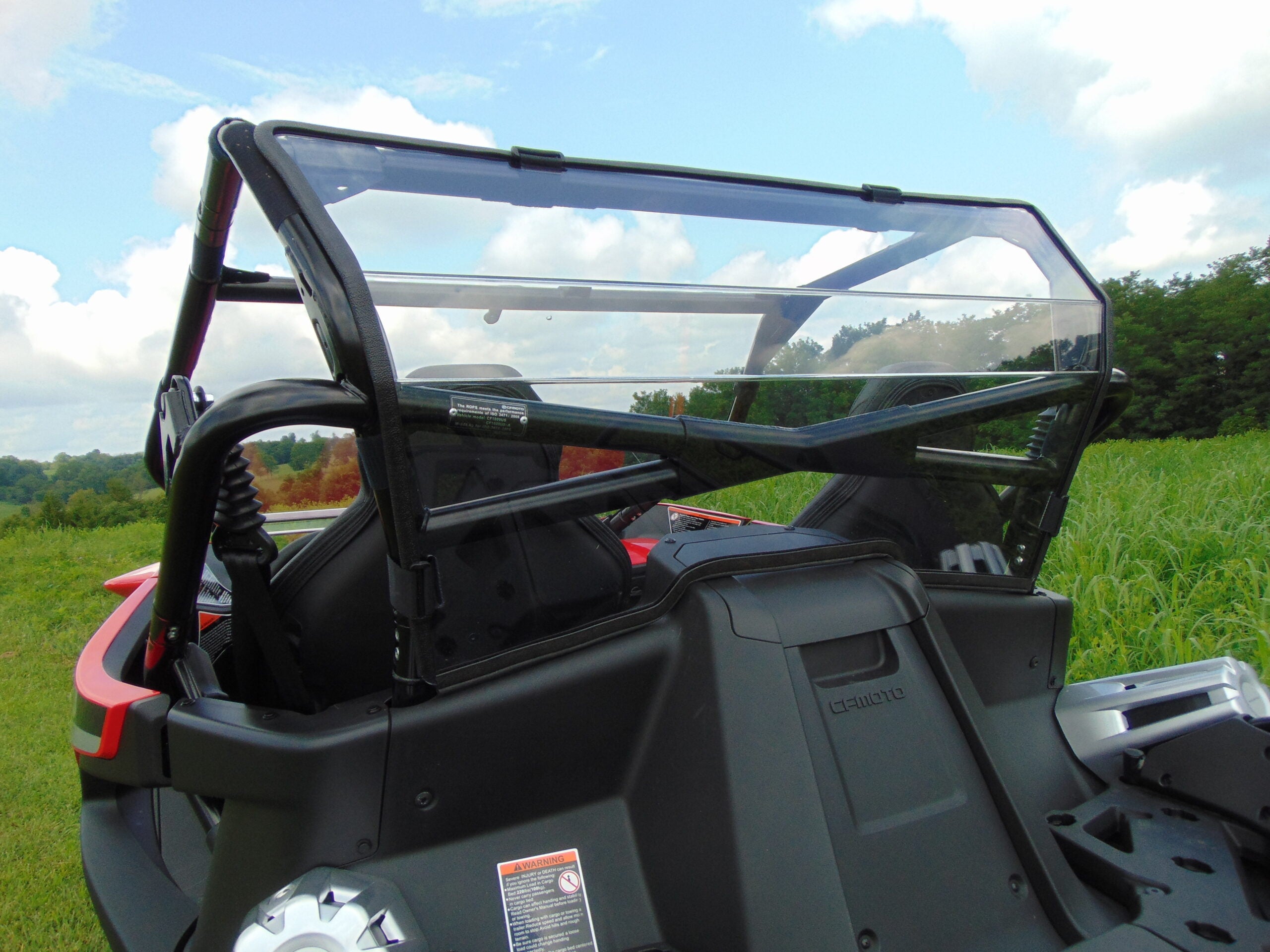 A close-up of the Cfmoto Zforce 950 Rear Windshield, with black seats visible through the clear, rectangular panel. The off-road vehicle is parked on green grass, and trees and a blue sky with clouds are in the background.