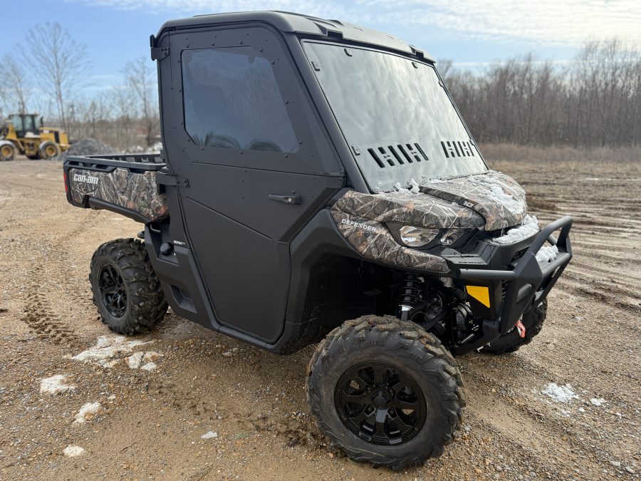 A Can Am Defender with a Full Aluminum Door Kit (converts from full to half, 2 door) is parked on a dirt road. It features off-road tires, tinted windows, and a closed cab; bare trees and construction equipment are in the background.