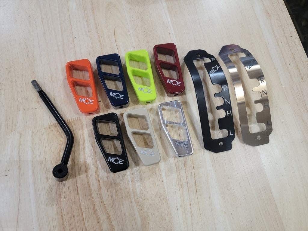 The Can-Am Defender HD8, HD9, or HD10 Ultimate Shifter Kit is displayed on light wood, featuring a black gear lever and colorful shift plates in red, orange, yellow, green, black, silver, clear, gray plus two metallic shift pattern plates.