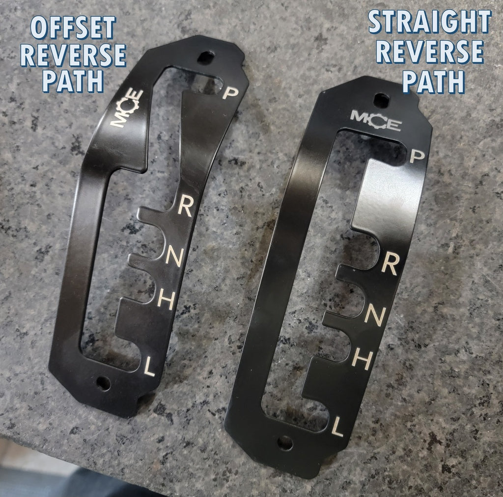 Shown are two black metal gate plates from the Can-Am Defender HD8, HD9, or HD10 Ultimate Shifter Kit. The left plate features an offset reverse path, while the right has a straight reverse path. Both are labeled P, R, N, H, and L.