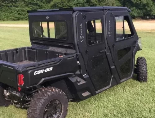 A black Can-Am Defender with large off-road tires and enclosed cabin doors features the Armor Tech Sliding Glass Rear Window. It sits on green grass, parked near trees and a dirt area, with a rear cargo bed visible.