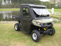 A green and black Armor Tech Can-Am Defender with a flip-up glass windshield, closed cab, off-road tires, and small cargo bed is parked on grass near a pond and trees—ideal for rugged outdoor or farm use.