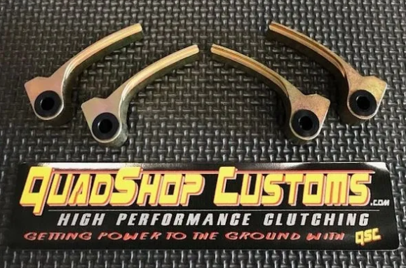 Four metallic clutch arms with black circular holes are arranged in an arc on a textured black surface. Below them is the Maverick Trail/Sport Clutch Kit sign from QuadShop Customs, stating HIGH PERFORMANCE CLUTCHING and GETTING POWER TO THE GROUND WITH Q