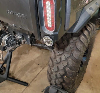 Close-up of a Can Am Defender featuring the Reverse Light Kit Plug and Play, highlighting the right taillight, round auxiliary light, and large treaded tire. The vehicle is on display in a garage with a black bag partially visible below.