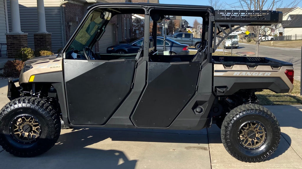 A 2019+ Polaris Ranger 1000 XP Crew fitted with Rival Half Doors, bronze wheels, off-road tires, and a rear metal rack is parked on a suburban driveway with houses, cars, and trees visible in the background.