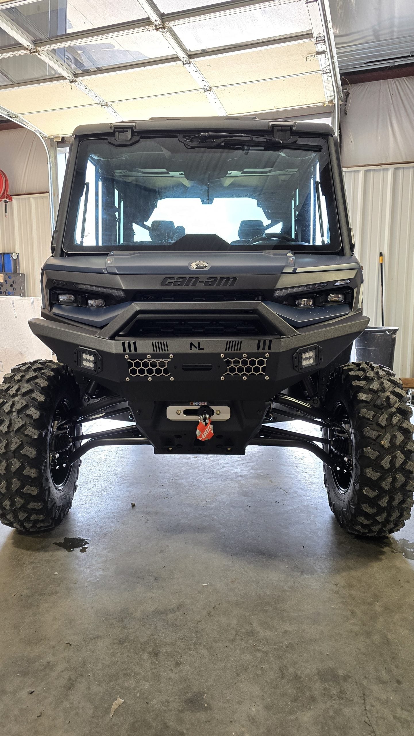 A 2026 Can-Am Defender HD11 with a front winch bumper and lights is parked in a garage with a concrete floor, roll-up door, corrugated metal walls, and tool shelves. The utility vehicle features rugged tires and tinted windows.