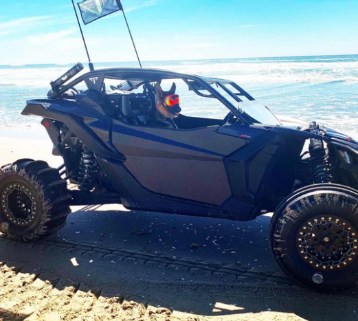 A dog with red goggles sits in the driver’s seat of a black dune buggy featuring Can-Am Maverick X3 Half Doors (2 Door) by Dirt Specialties, parked on a sandy beach near the ocean with a flag and deep tire tracks in the sand.
