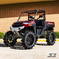 A Polaris Ranger with an S3 Power Sports 8 Lift Kit (2018+ Body Style), featuring large mud tires, a roll cage, black seats, and rugged suspension, is parked on pavement with a brick building and green bushes in the background.