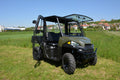 A Polaris Ranger XP570/SP Mid-Size Cab Kit (2022-Present) in green, featuring large tires, open side doors, and a transparent roof, is parked on grass with houses and trees in the background under a clear blue sky. The vehicle faces slightly left.
