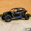 A black Can-Am Maverick X3 equipped with S3 Power Sports 2017+ Nerf Bars and blue accents is parked on concrete in front of a tan brick wall and green bushes. Large rugged tires, roll bars, an open cabin, and the SX logo are visible.