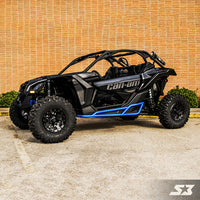 A black Can-Am Maverick X3 equipped with S3 Power Sports 2017+ Nerf Bars and blue accents is parked on concrete in front of a tan brick wall and green bushes. Large rugged tires, roll bars, an open cabin, and the SX logo are visible.