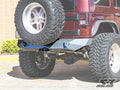 A red Jeep Wrangler JK fitted with the S3 Power Sports Ambush Series rear bumper and large rugged tires is parked on concrete near a brick wall, with its spare tire mounted on the back and green shrubs to the left.