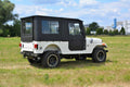 A white ROXOR4 with a black Full Hard Enclosure Cab Kit by Hardcabs is parked on grass. The Jeep-style SUV features rugged tires, side mirrors, and rectangular taillights, with trees, a fence, and industrial buildings in the background.