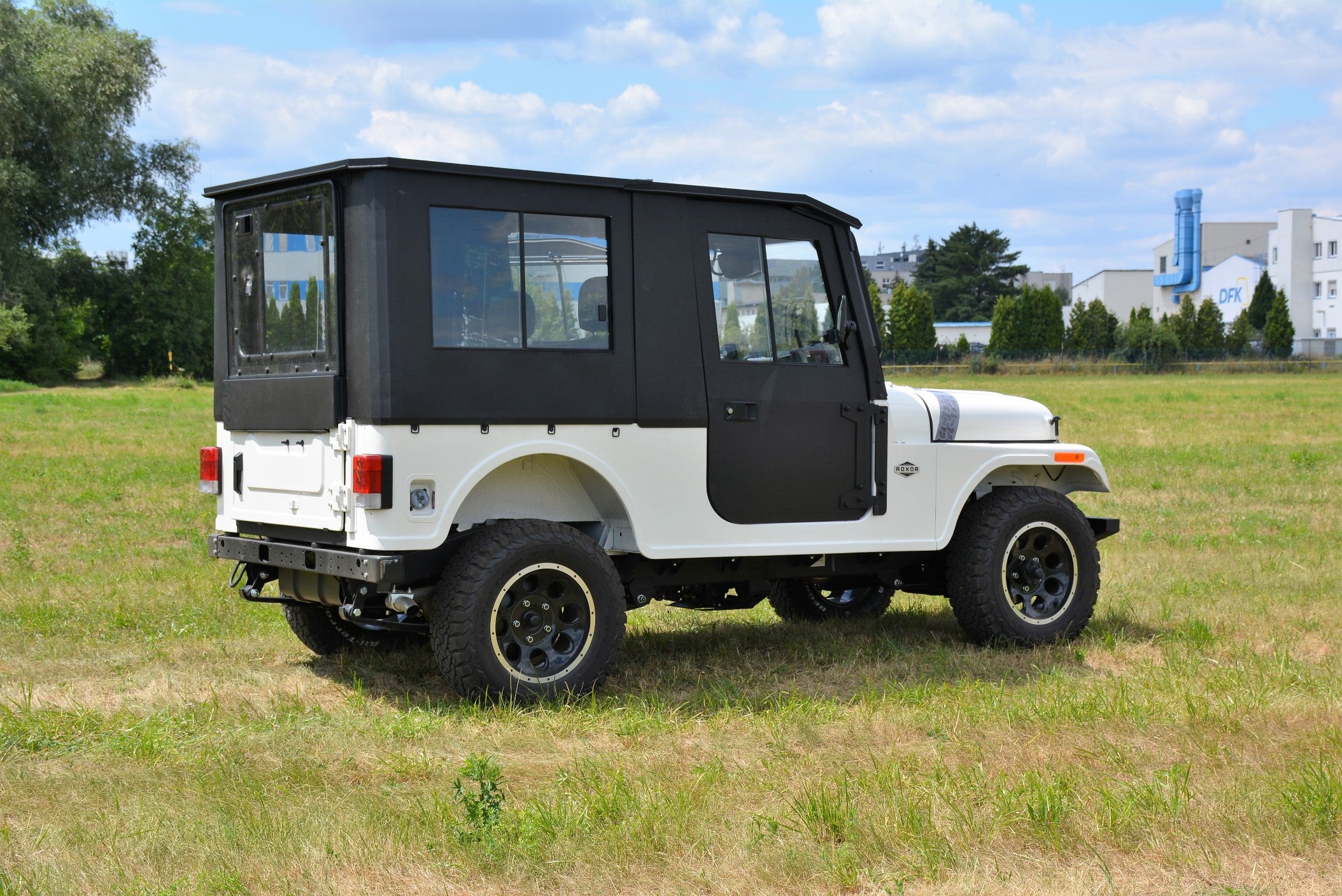 A white ROXOR4 with a black Full Hard Enclosure Cab Kit by Hardcabs is parked on grass. The Jeep-style SUV features rugged tires, side mirrors, and rectangular taillights, with trees, a fence, and industrial buildings in the background.