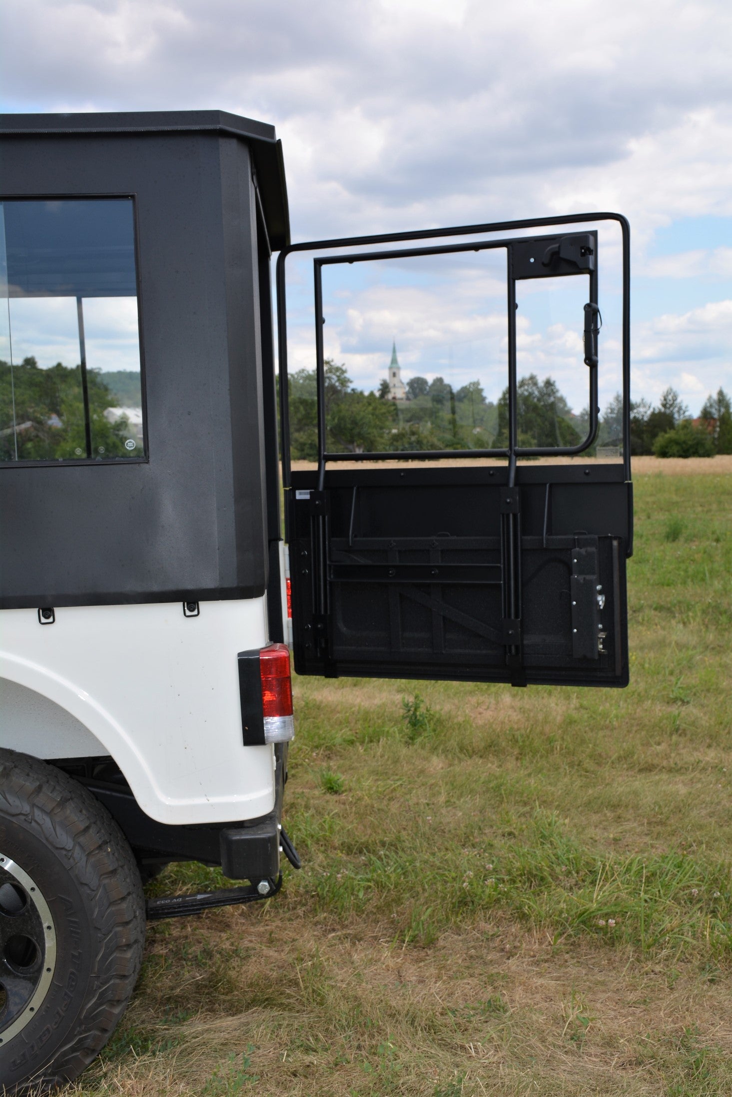 A ROXOR with the ROXOR4 Full Hard Enclosure Cab Kit by Hardcabs is parked on grass with its rear door open, beneath a partly cloudy sky. Distant trees and a building with a tall spire can be seen in the background.