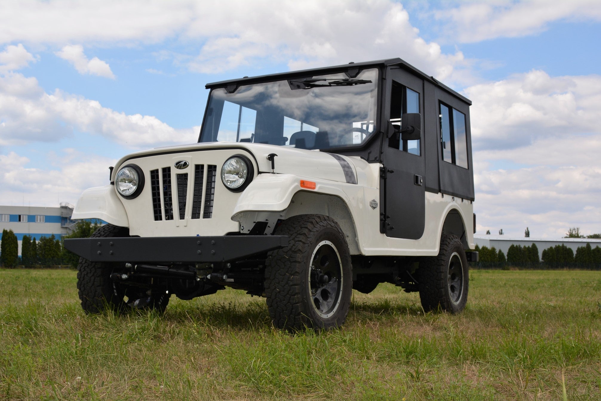 A ROXOR4 SUV equipped with the Full Hard Enclosure Cab Kit by Hardcabs, featuring a boxy black hardtop and rugged tires, is parked on grass under a partly cloudy sky. Buildings and shrubs are visible in the background.