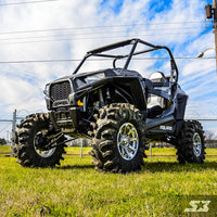 A Polaris General with an S3 Power Sports 3 Bracket Lift Kit and rugged tires sits on grass, viewed low with blue sky, power lines, and a wire fence behind. The “SX” logo appears at the lower right.