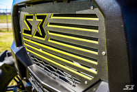 Close-up of the S3 Power Sports 2014-2017 Polaris Rzr S3 Nation Grille in black metal with yellow accents, a stylized “S3” logo and star, horizontal slats, and angular lines. Sunlight reflects on it; blurry grass and fence are in the background.