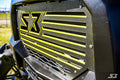 Close-up of the S3 Power Sports 2014-2017 Polaris Rzr S3 Nation Grille in black metal with yellow accents, a stylized “S3” logo and star, horizontal slats, and angular lines. Sunlight reflects on it; blurry grass and fence are in the background.