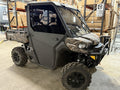 A CanAm Defender with a Half Aluminum Door Kit, black and camouflage, is parked on a concrete floor indoors. It features large rugged tires, two side doors, a front windshield, and a small cargo bed amid warehouse shelves and boxes.