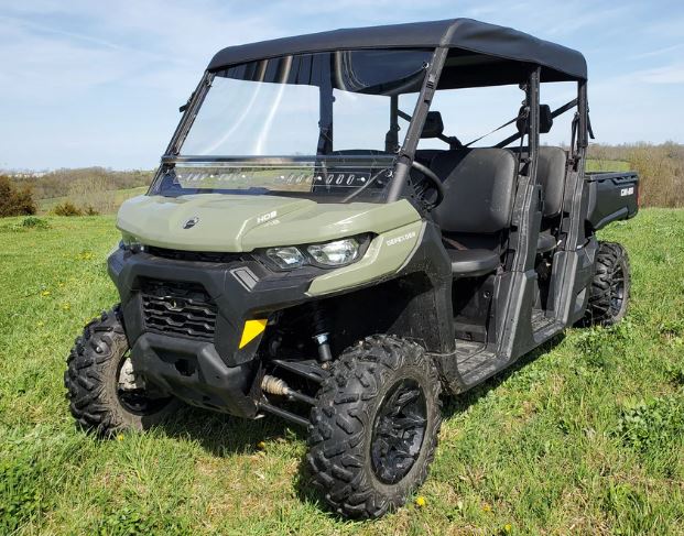 A green Can-Am Defender Max 4 - Soft Top UTV with black wheels, a soft roof, off-road tires, open sides, and seating for multiple passengers is parked on grass in a field with trees and blue sky in the background.