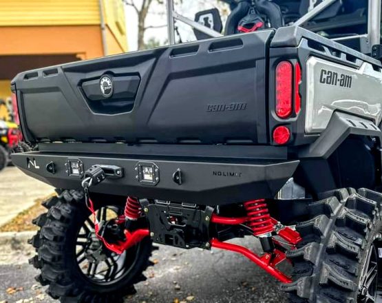 A close-up shows the Can-Am Defender Rear Winch Bumper with Lights, featuring red springs, rugged black tires, a custom black bumper with integrated lights and winch. The tailgate and Can-Am branding are clearly visible.