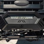 Close-up of the 2011-2016 Ford F-250 Spartan Front Bumper by Road Armor. This textured black, sheet metal pre-runner guard features a center logo, built-in LED light bar, angular lines, and visible suspension and tire components.