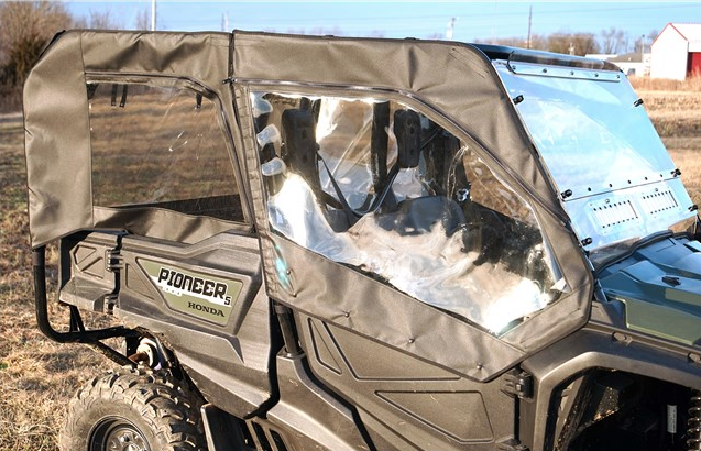 A Honda Pioneer 1000-5 with a Front/Back Soft Door Kit and Middle/Rear Windows, featuring black fabric and clear panels for full cab weather protection, is parked outdoors on dry grass with trees and a red barn in the background.