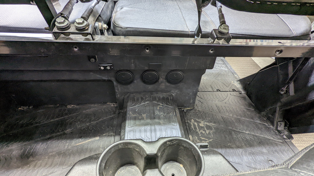 The image shows the rear floor area of a Polaris Ranger XP1000 Crew (2018-2024), featuring a black textured rubber mat, three round vents on the console, two cup holders in front, plus a fabric seat back and metal brackets.