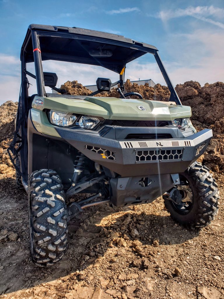 A Can-Am Defender with a Front Gen 2 Bumper is parked on uneven dirt terrain. Its large, rugged tires and the clear sky with brown soil mounds in the background highlight its off-road capability.