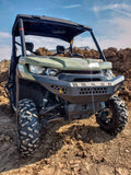A Can-Am Defender with a Front Gen 2 Bumper is parked on uneven dirt terrain. Its large, rugged tires and the clear sky with brown soil mounds in the background highlight its off-road capability.