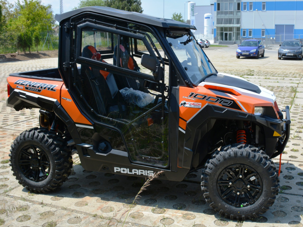 A Polaris General 1000 Hard Cab Enclosure in black and orange is parked outdoors on patterned concrete, featuring rugged tires, clear side doors, visible suspension, and a modern blue-accented building in the background.