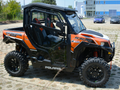 A Polaris General 1000 Hard Cab Enclosure in black and orange is parked outdoors on patterned concrete, featuring rugged tires, clear side doors, visible suspension, and a modern blue-accented building in the background.