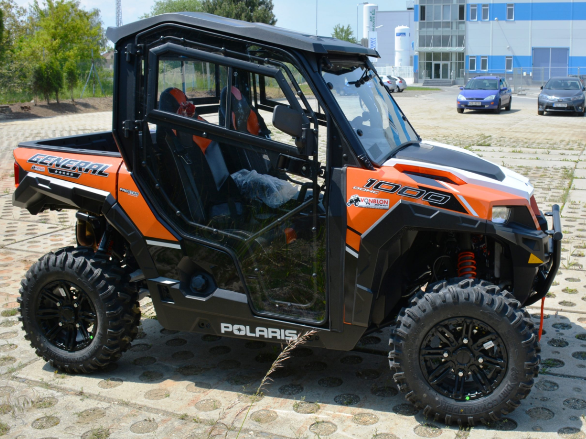 A Polaris General 1000 Hard Cab Enclosure in black and orange is parked outdoors on patterned concrete, featuring rugged tires, clear side doors, visible suspension, and a modern blue-accented building in the background.