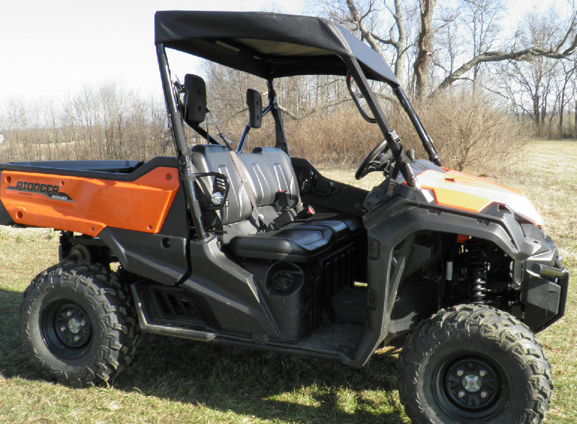 A Honda Pioneer 700 (2014+) Soft Top UTV with large rugged tires, two black seats, a roll cage, and no doors is parked on grass. Leafless trees and brown shrubs are in the background under a clear sky.