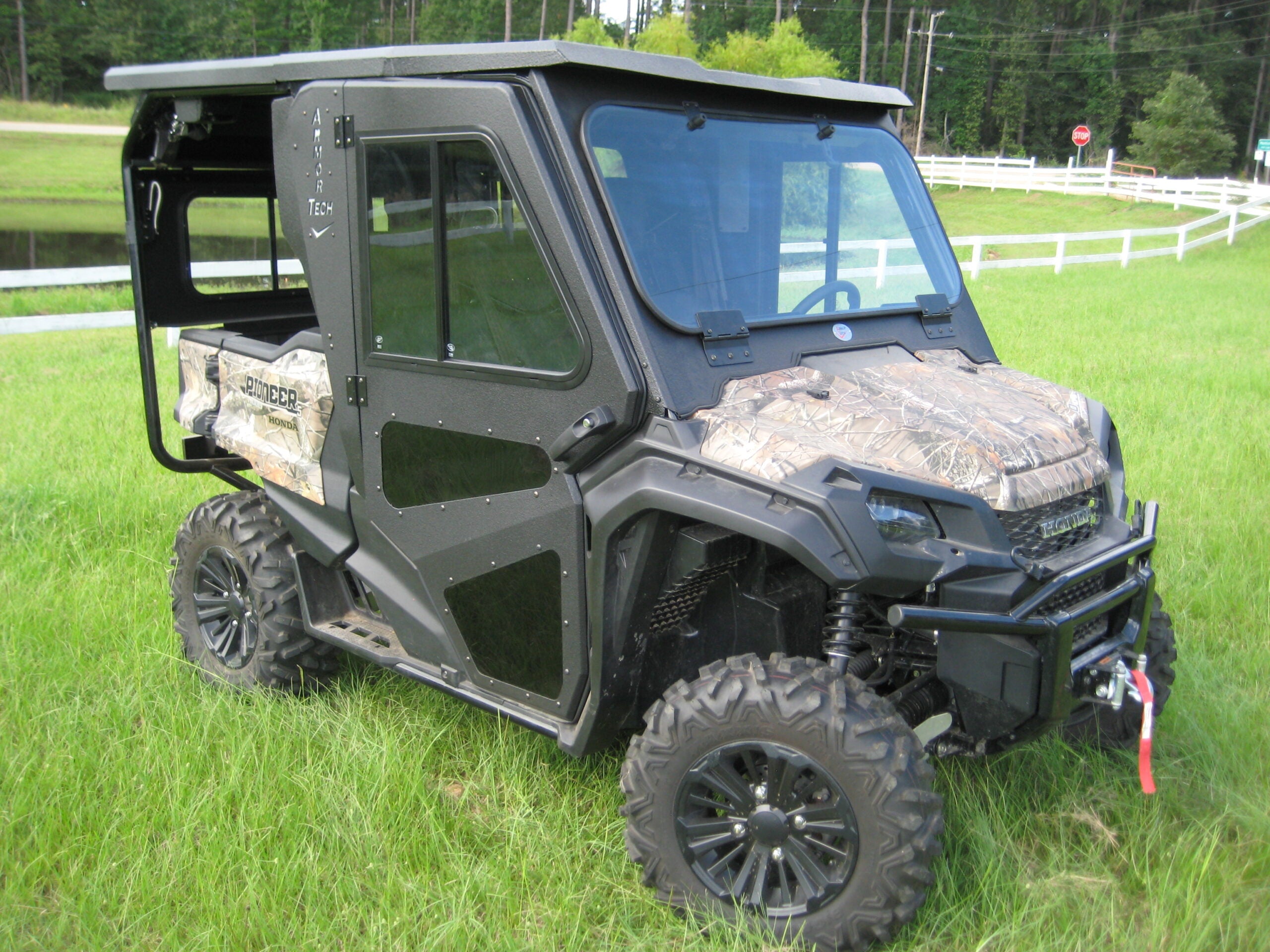 A Honda Pioneer 1000-5 Full Hard Cab Enclosure by Armor Tech, featuring black and camouflage accents, is parked on grass near a white fence. The closed cab UTV has large windows, rugged tires for off-road use, with trees and power lines behind.