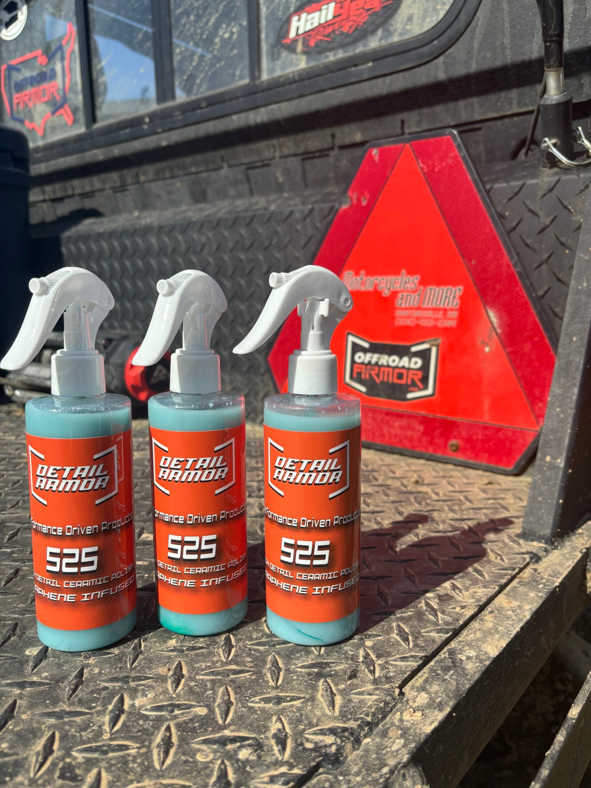 Three bottles of Detail Armor 525 High Detail Ceramic Polish Infused with Graphene rest on a textured black truck bed beside a red Offroad Armor toolbox, highlighted by sunlight and casting distinctive shadows.