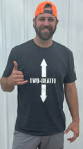 A bearded man in a neon orange backward cap and gray shorts stands by a metal wall, giving a hang loose gesture. He wears the Two Seater - Offroad Armor Next Level T-Shirt with bold TWO-SEATER text and large white arrows.