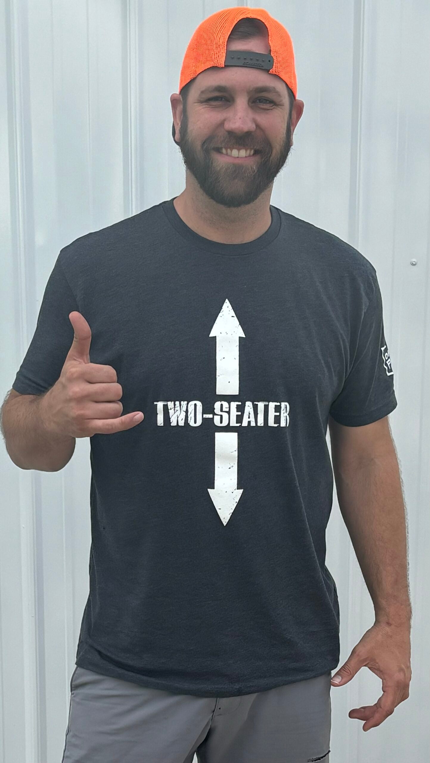 A bearded man in a neon orange backward cap and gray shorts stands by a metal wall, giving a hang loose gesture. He wears the Two Seater - Offroad Armor Next Level T-Shirt with bold TWO-SEATER text and large white arrows.