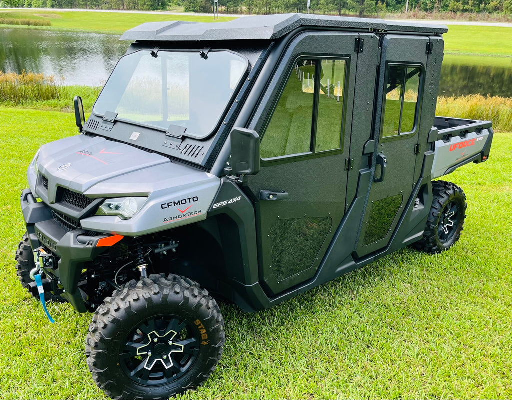 A CFMOTO UFORCE 1000XL with Armor Tech Full Steel Doors and sliding windows is parked on green grass near a pond. The black side-by-side utility vehicle has rugged tires, tinted windows, decals, and a front bumper winch.