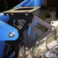 Close-up of an S3 Power Sports 2017+ Can-Am Maverick X3 Transmission Brace, featuring a black metal bracket with triangular cutouts mounted to the blue chassis and silver engine casing with bolts. Parts look new, with a blurred workshop background.
