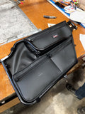 A pair of Dirt Specialties Can-Am Maverick X3 Door Bags with pockets, zippers, and a GRT logo are on a wooden workbench surrounded by tools and materials, with a sneaker-clad foot visible on the right edge of the image.