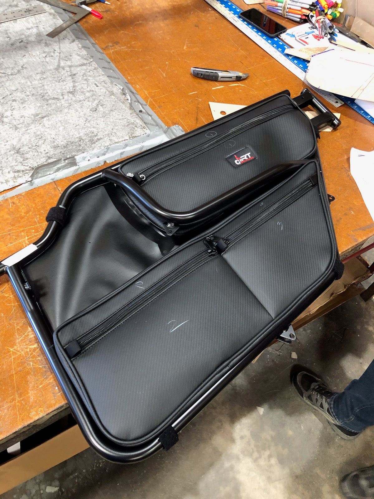 A pair of Dirt Specialties Can-Am Maverick X3 Door Bags with pockets, zippers, and a GRT logo are on a wooden workbench surrounded by tools and materials, with a sneaker-clad foot visible on the right edge of the image.